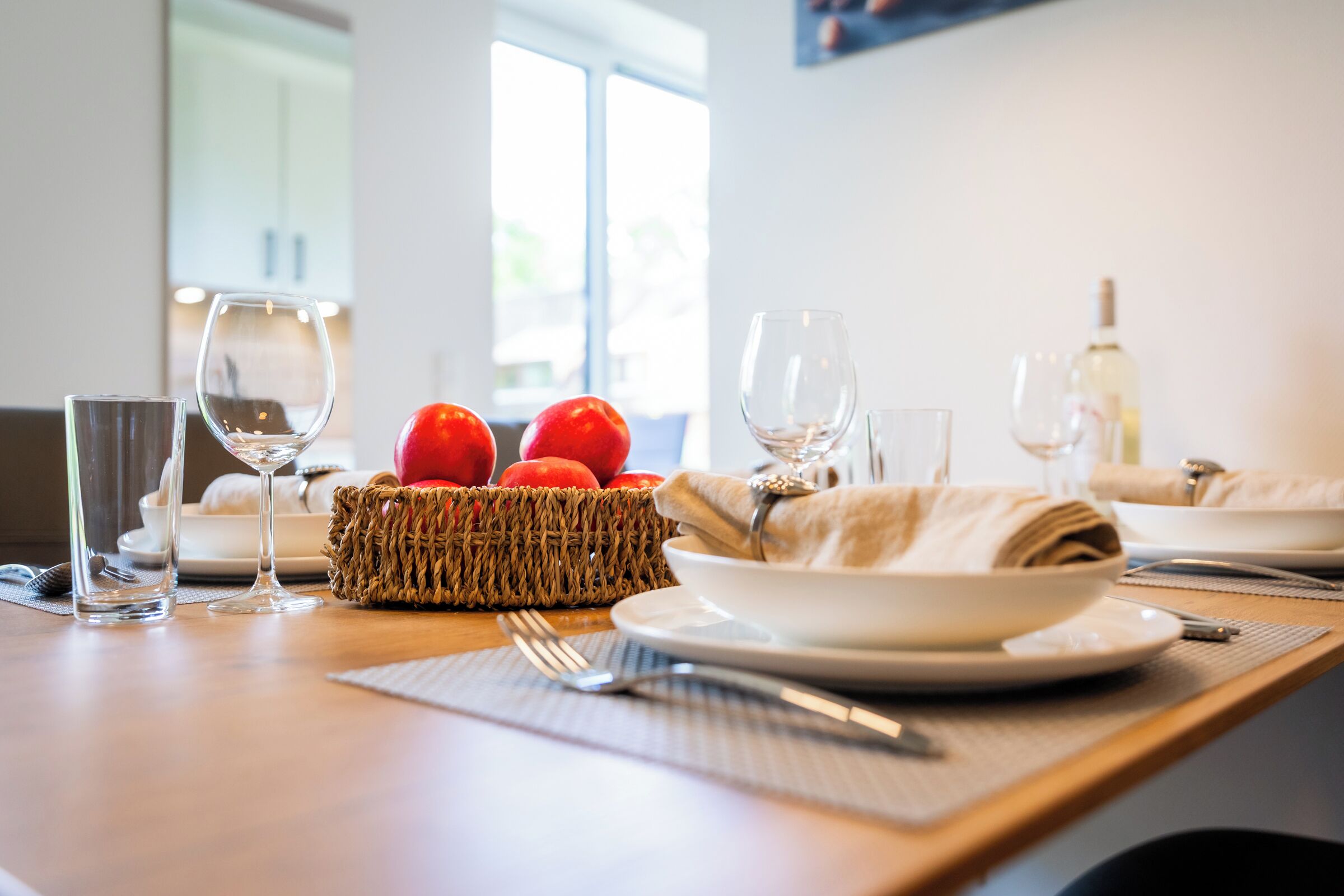 A dining table set with plates, glasses, cutlery, a woven basket with red apples, and a bottle of white wine.