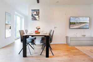 A modern dining area with a wooden table, black chairs, and a wall-mounted TV displaying a beach scene. - Grömitz WindStärke 7 - Apartment 1. Ocean view. Sauna. Patio (Grömitz)