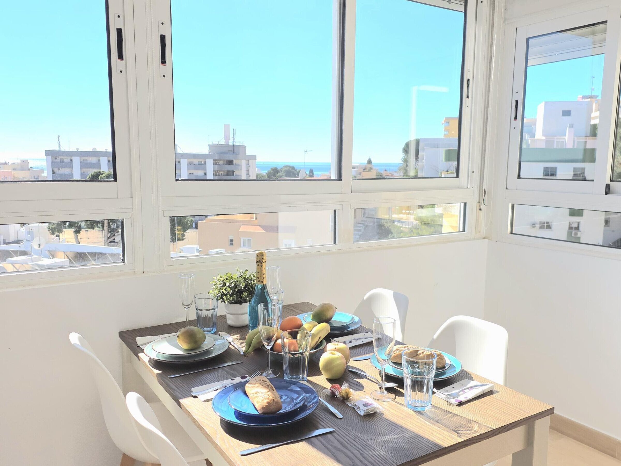 Comedor con mesa para dos, una planta en maceta y vistas al mar a través de grandes ventanales.