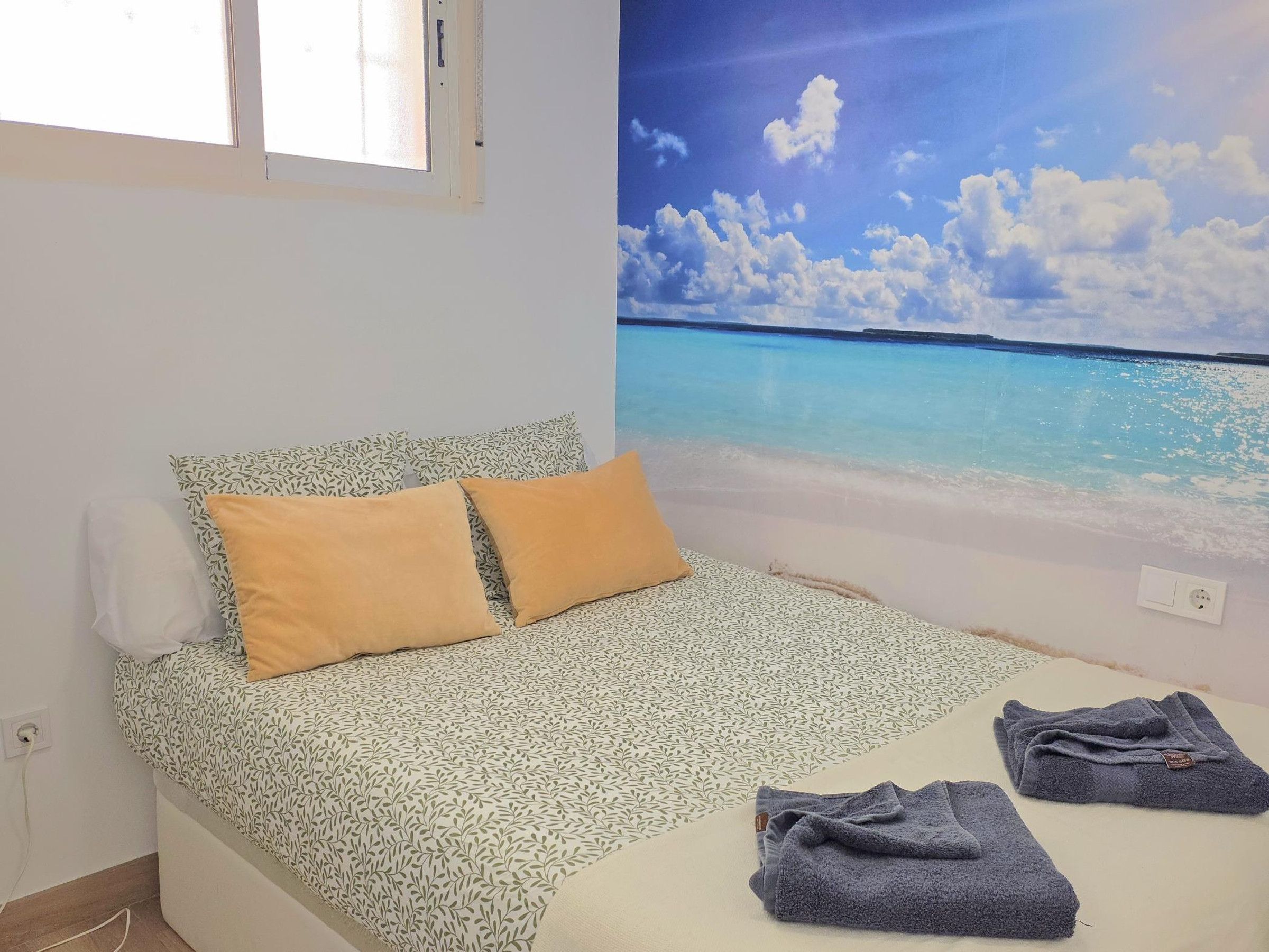 A bed with a patterned bedspread, two pillows, and a view of a beach through a wall mural.