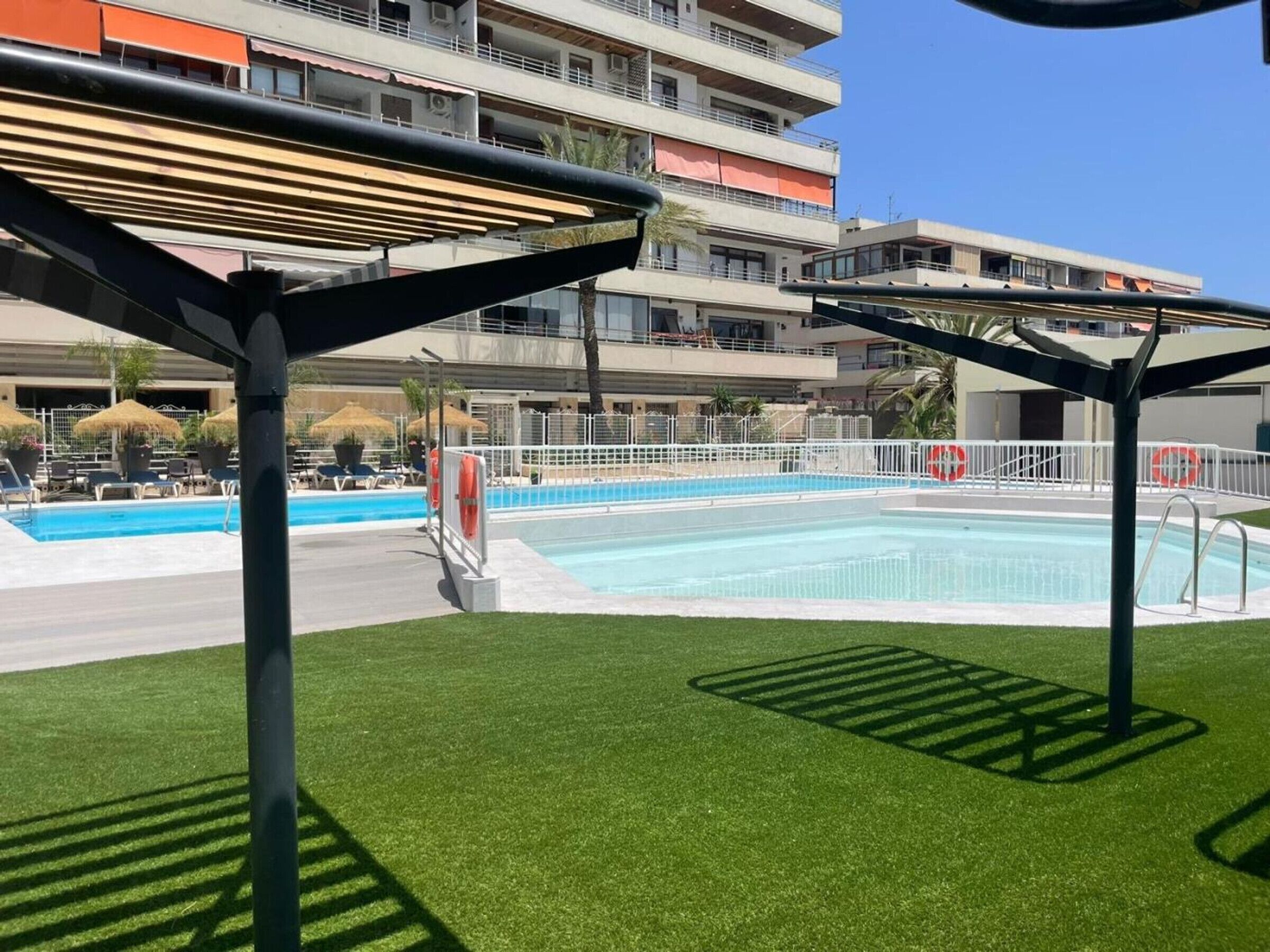 Una piscina con un borde perimetral, un puesto de socorrista y un edificio con balcones al fondo.