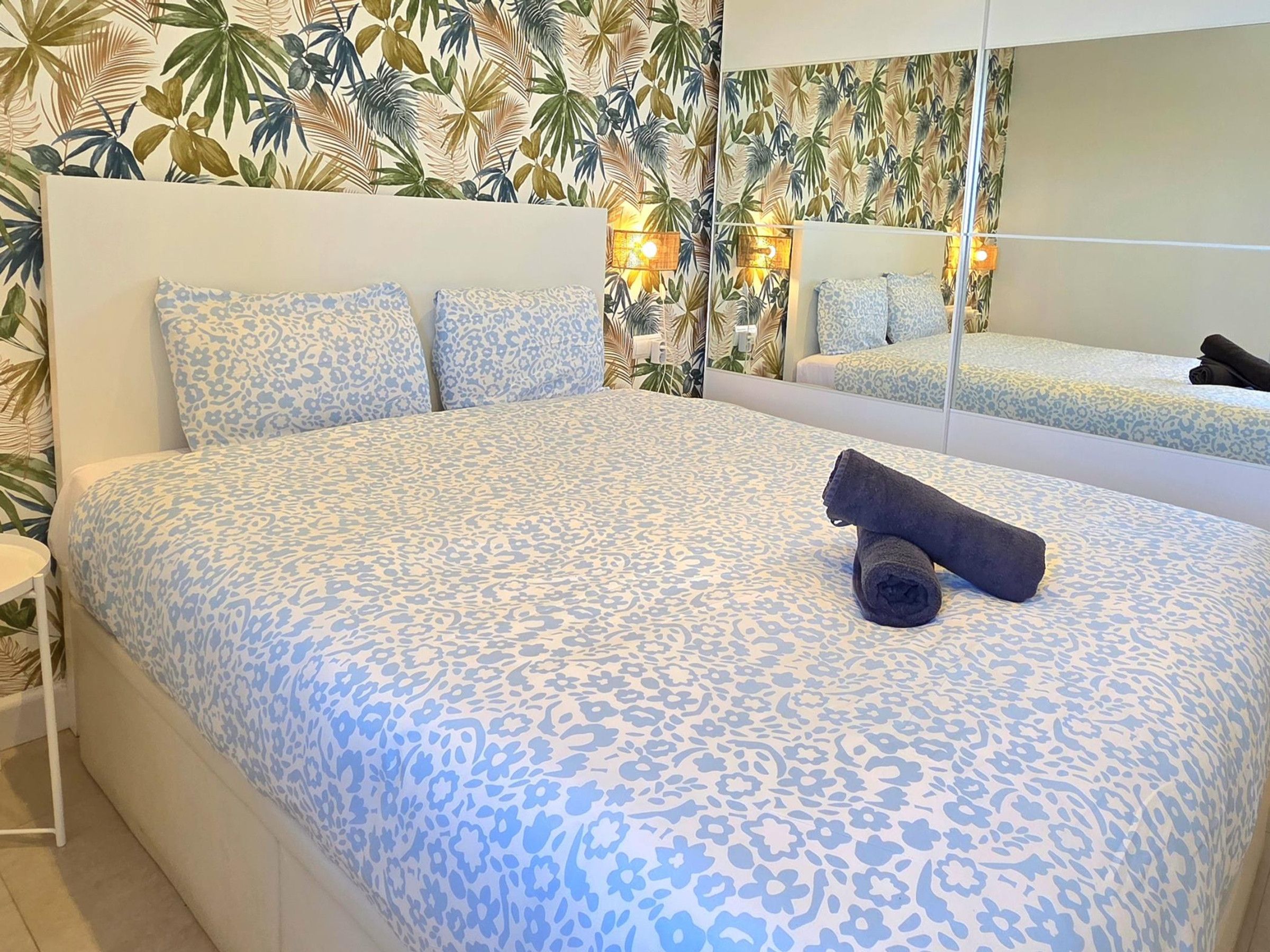 Hotellihuone, jossa on sänky, yöpöytä, peili ja trooppikuviollinen tapetti.