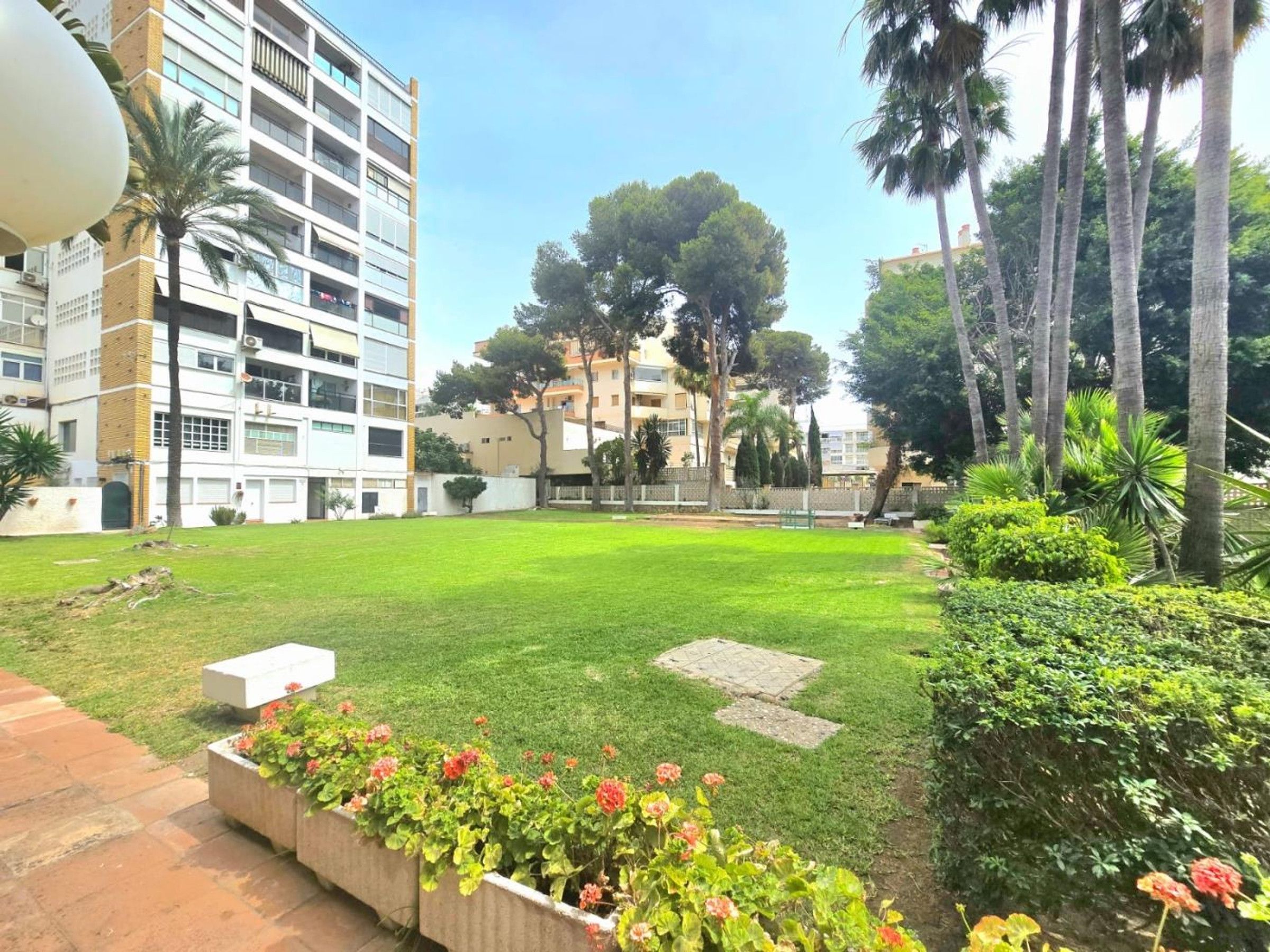 Un patio con césped, una jardinera de concreto con flores y un edificio de varios pisos y balcones.