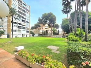 Un patio con césped, una jardinera de concreto con flores y un edificio de varios pisos y balcones.