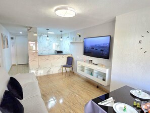 A modern living room with a flat-screen TV, a dining table set for two, and a kitchen area with marble countertops. - Central apartment with pool - 4 guests (Torremolinos)