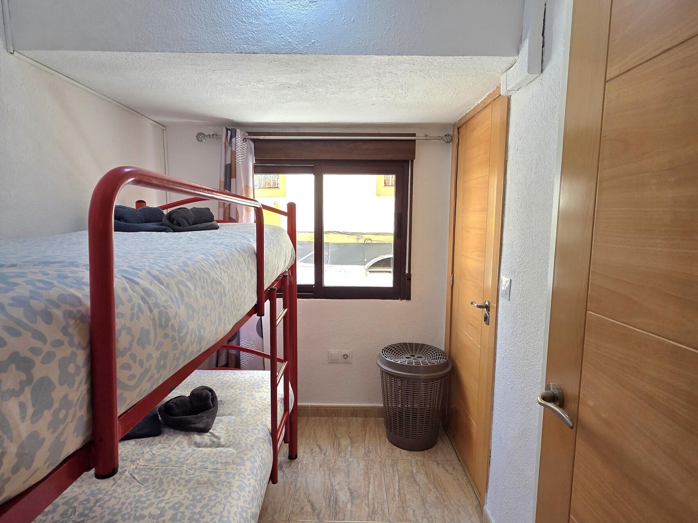 Um quarto pequeno com uma cama de solteiro, uma janela, uma porta e uma lixeira.
