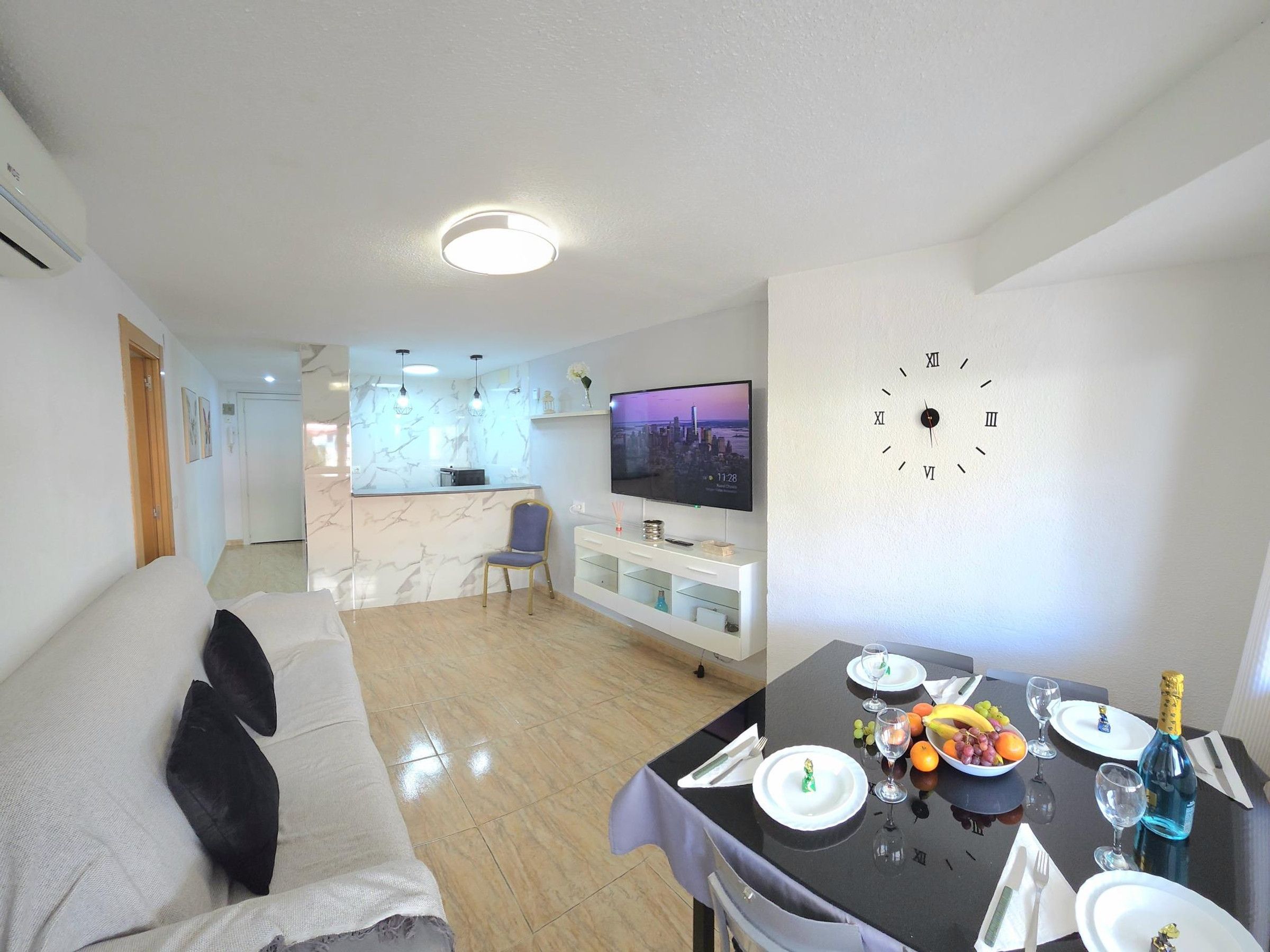 Ein modernes Wohnzimmer mit einer Couch, einem Esstisch für zwei Personen, einem Fernseher und einer Wanduhr.