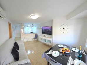 A modern living room with a sofa, dining table set for two, a TV, and a wall clock. - Central apartment with pool - 4 guests (Torremolinos)