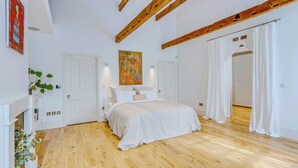 A bedroom with a wooden floor, white walls, a bed with white bedding, a painting above the bed, and a door leading to another room. - Luxury London Six-Bedroom House in Marylebone - Moments from Regent’s  Park (London)