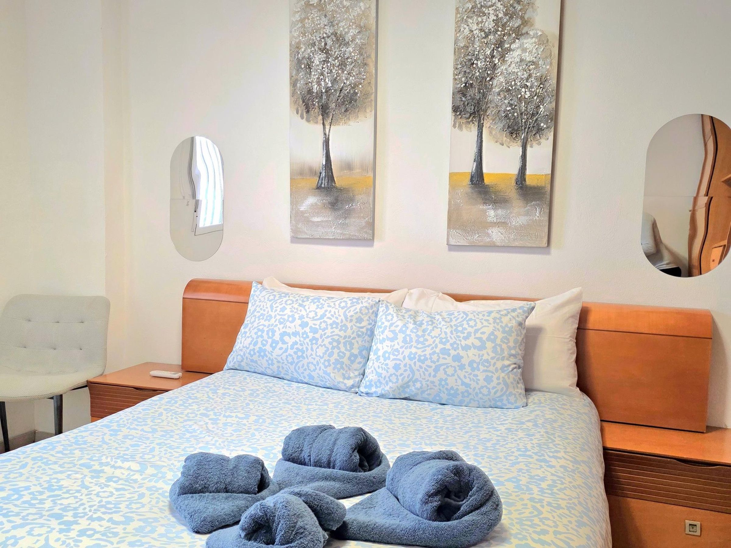 A hotel room with a bed, two wall-mounted paintings, a mirror, and a chair.
