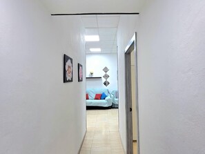 Ein Flur mit einer Tür rechts, einem hellen Boden und einem Raum im Hintergrund mit einem Bett und Kissen.