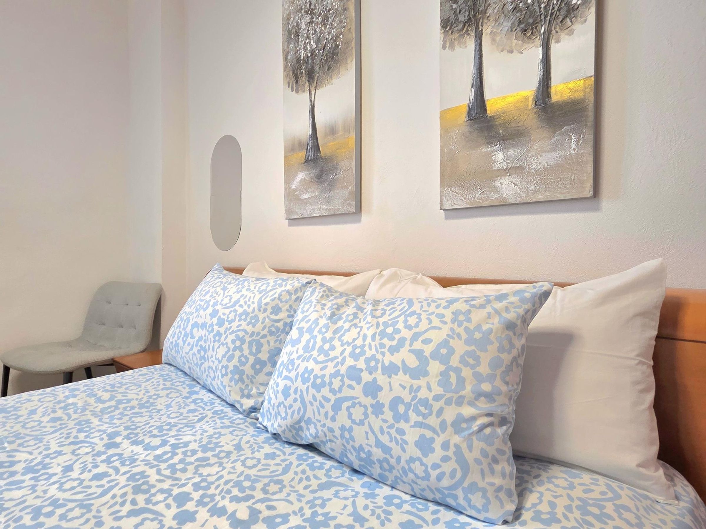A bedroom with a bed, two pillows, a chair, and two paintings on the wall.