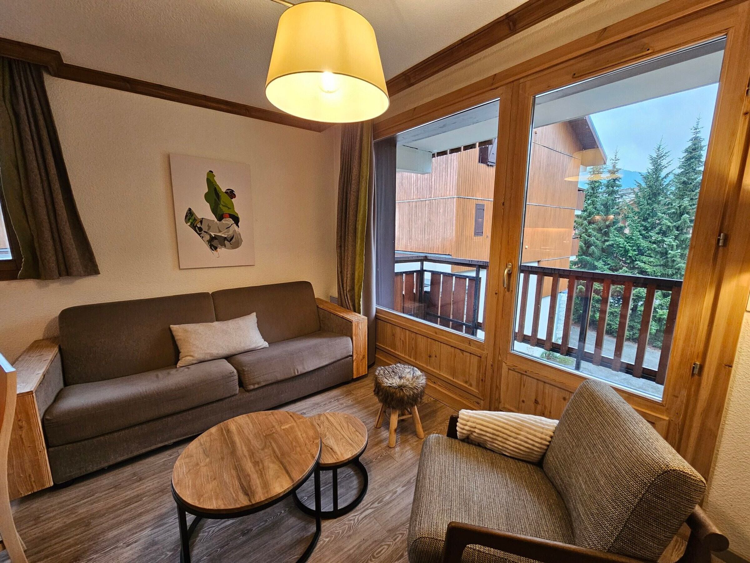 A cozy living room with a sofa, two wooden tables, a small stool, and a large window with a balcony view.