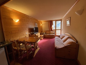 Ein gemütliches Zimmer mit einer Holzwand, einem Fernseher, einem Sofa und einem Esstisch mit Stühlen.