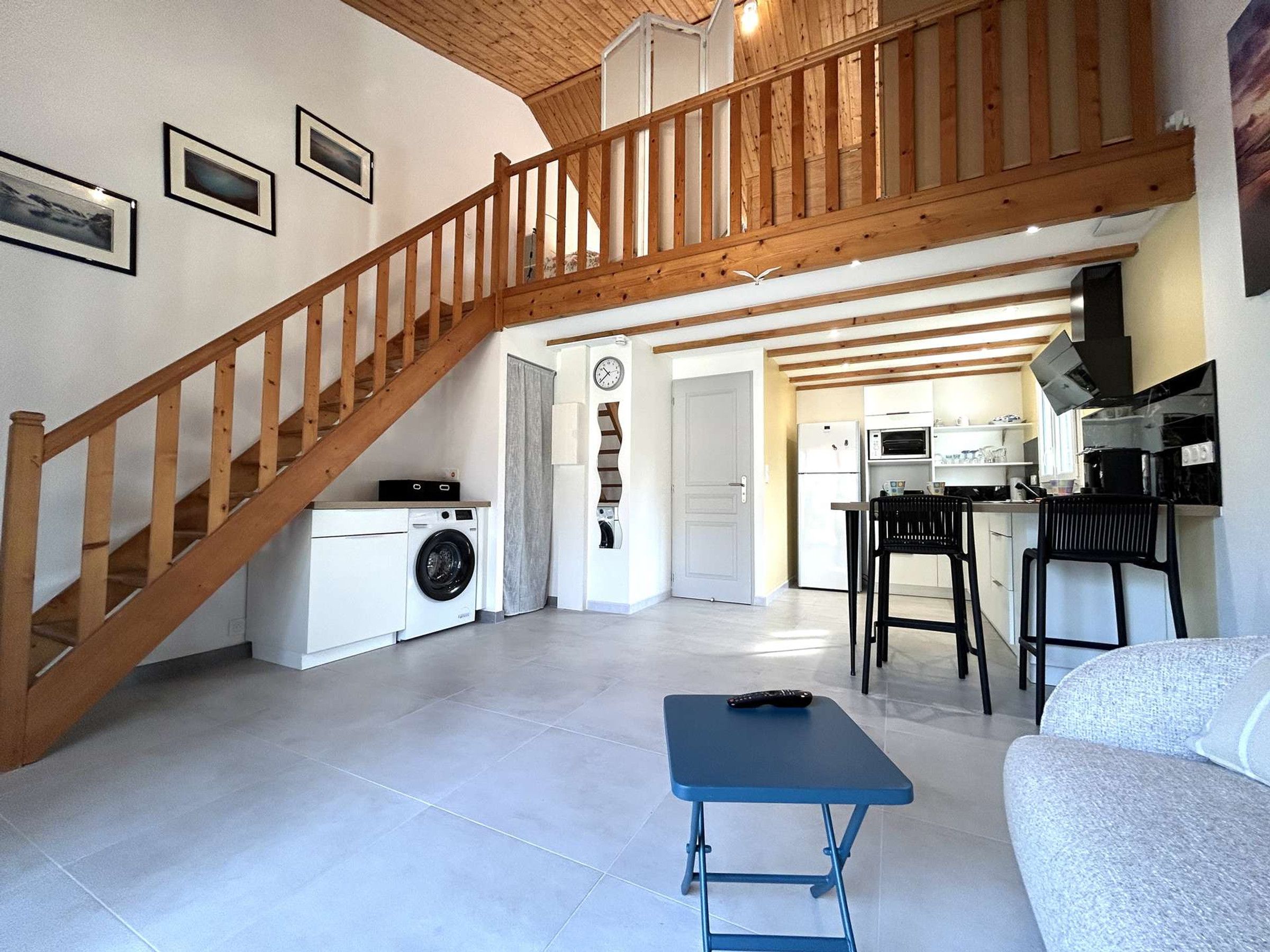 A spacious living area with a wooden staircase, a washing machine, a kitchen with modern appliances, and a blue table.