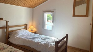Makuuhuoneessa on puinen katto, yksi sänky valkoisilla vuodevaatteilla, yöpöytä lampun kanssa ja ikkuna, josta avautuu näkymä vehreään maisemaan.