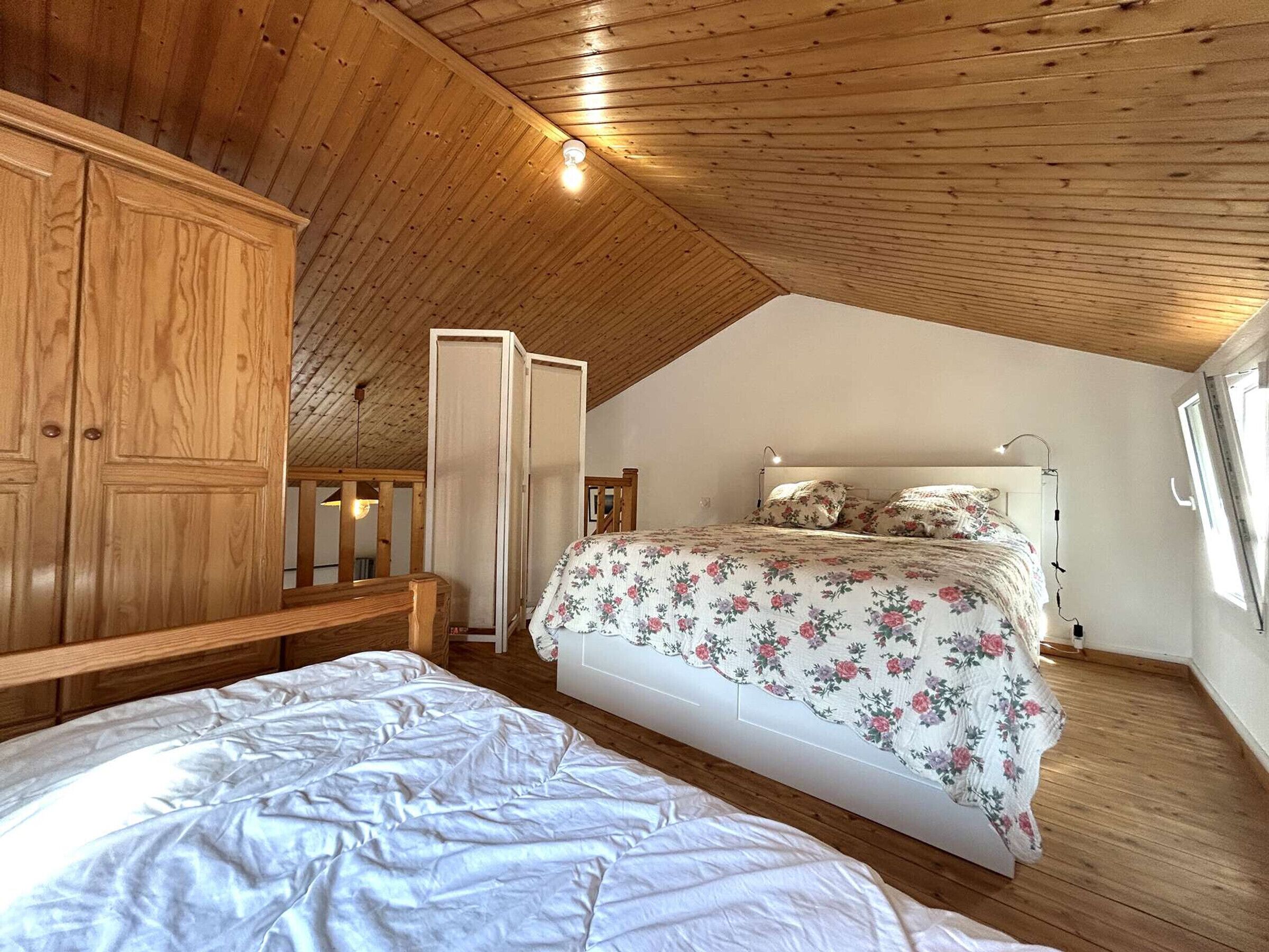 A bedroom with a sloped ceiling, wooden wardrobe, a bed with floral bedding, and a bedside table.