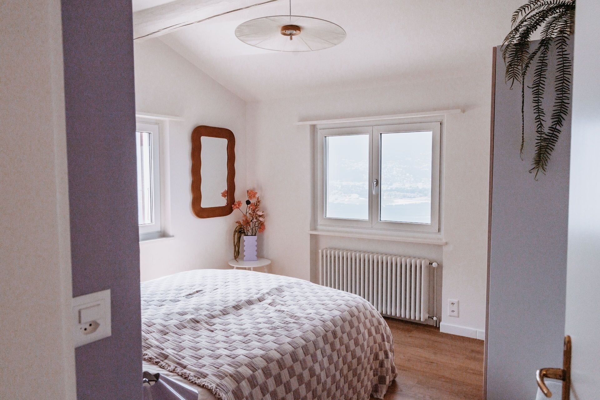 Een slaapkamer met een bed, een raam met uitzicht, een radiator en een spiegel aan de muur.