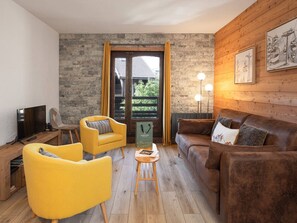 A living room with a stone wall, wooden paneling, a brown leather sofa, yellow armchairs, a TV, and a table with a book. - Pleasant apartment, mountain view - 4 guests (Le Monêtier-les-Bains)