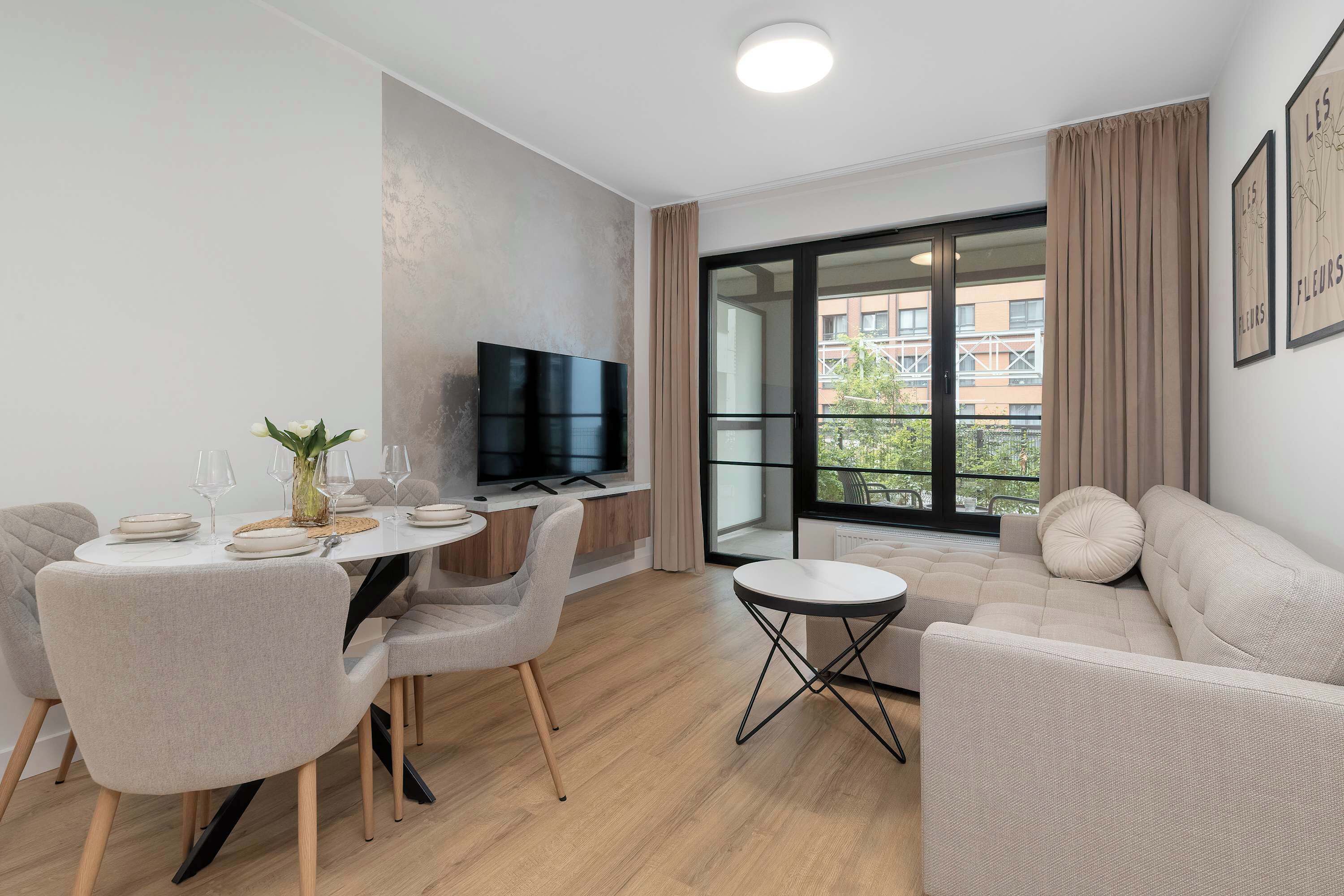 A modern living room with a dining area, a flat-screen TV mounted on the wall, and a large window offering a view of the city.