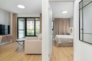 En modern lägenhet med en platt-TV, en soffa, ett litet bord, en säng med ett vitt täcke och ett badrum med en spegel och belysning.