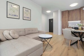A modern living room with a sectional sofa, a small round table, and a dining area with a wooden cabinet and a glass table. - Comfortable Apartment with a Garden | Parking (Gdańsk)