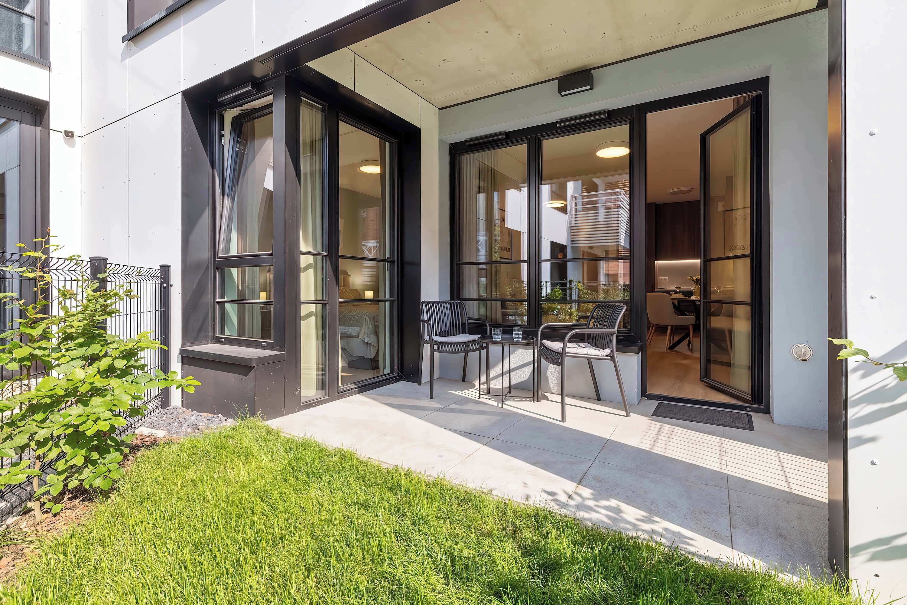 A modern building with a glass door entrance, a small outdoor seating area with two chairs and a table, and a well-maintained grassy area.