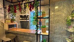 A kitchen area with a microwave, bar stools, a small table, and shelves with plants and bottles.