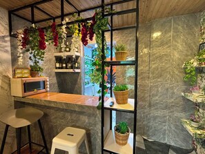 A kitchen area with a microwave, bar stools, a small table, and shelves with plants and bottles. - Ian's Place (Bacolod)
