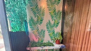 A narrow outdoor space with a wall decorated with a leaf pattern, a small table with a potted plant, a black trash bin, and a broom.