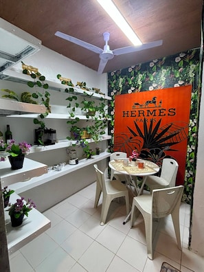 Un comedor con una mesa redonda, sillas blancas y un ventilador de techo. Se observan estantes con plantas y un cartel que dice 'HERMES PARIS'.