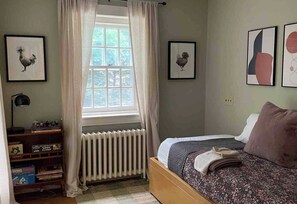 Un dormitori amb un llit, un radiador, una finestra amb cortines, un escriptori amb llibres i quadres al mur.