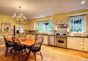 Une cuisine avec une table à manger, des chaises, un lustre et une fenêtre ornée de pots suspendus.