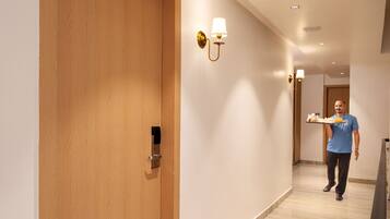 Corredor de hotel com porta de madeira, luminária na parede e uma pessoa carregando uma bandeja.