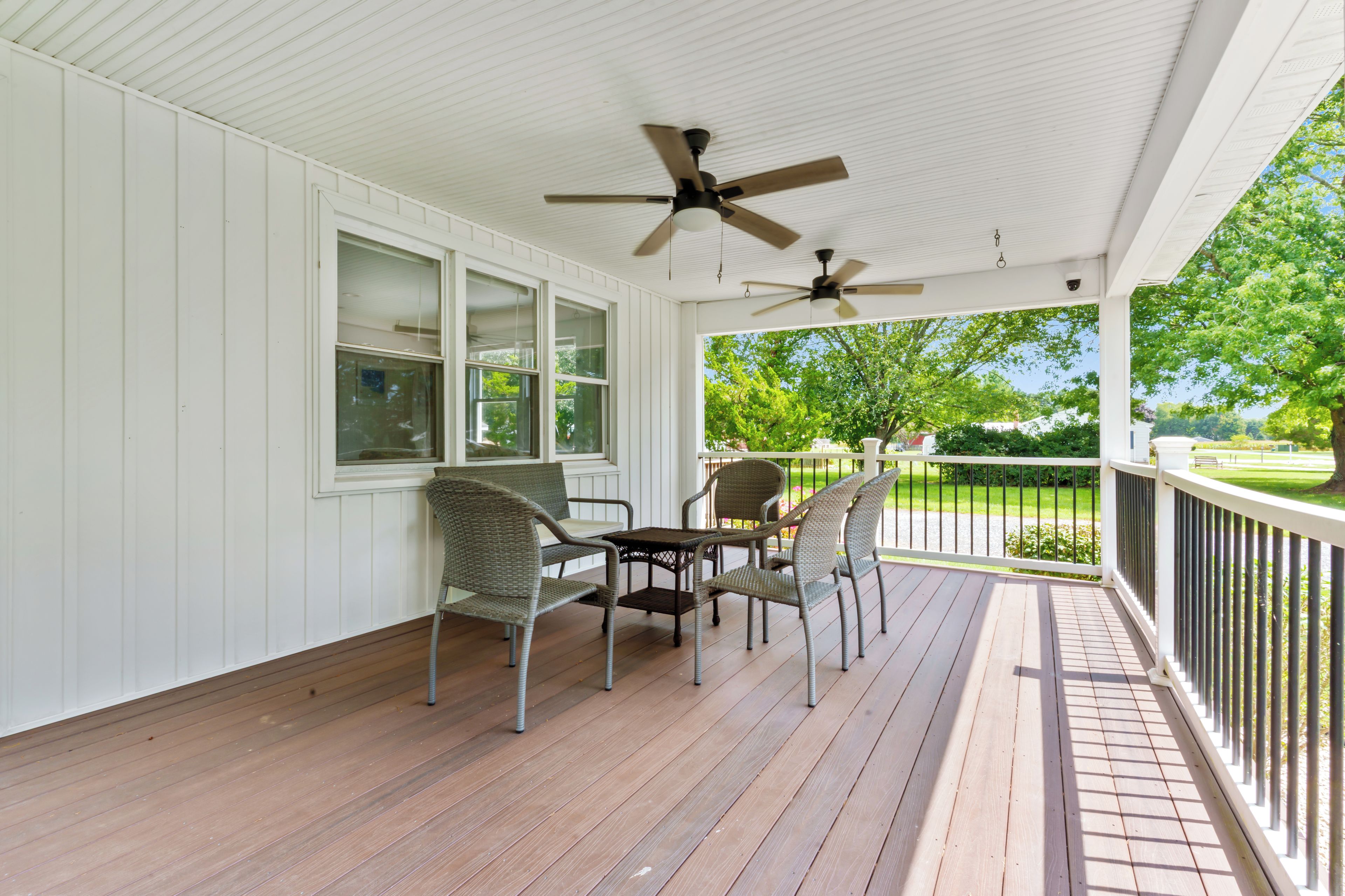 Une véranda couverte avec un ventilateur de plafond, un plancher en bois et des chaises en osier.