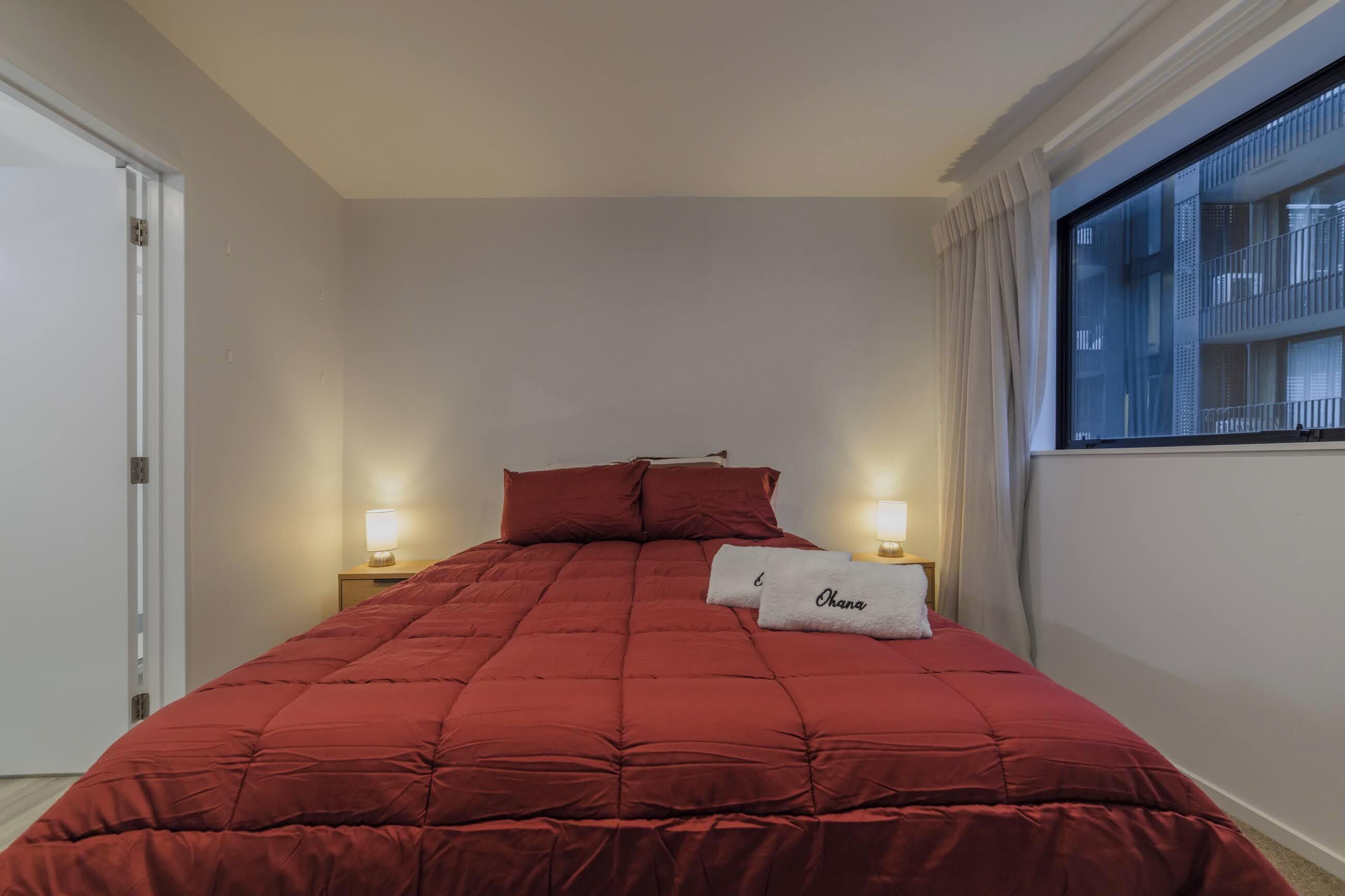 Una habitación de hotel con una colcha roja, dos lámparas de noche, un ventanal con cortinas y una puerta.