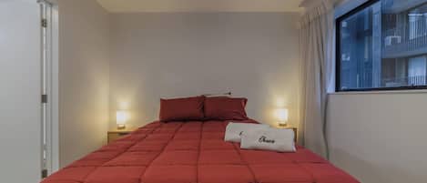 Una habitación de hotel con una colcha roja, dos lámparas de noche, un ventanal con cortinas y una puerta.