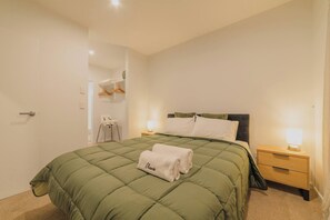 A neatly made bed with a green quilt, two bedside lamps, a small wooden nightstand, and a white towel with the name 'Chana' on it. - Modern Christchurch 4BR w/ Balcony Near NZ Stadium (Christchurch)