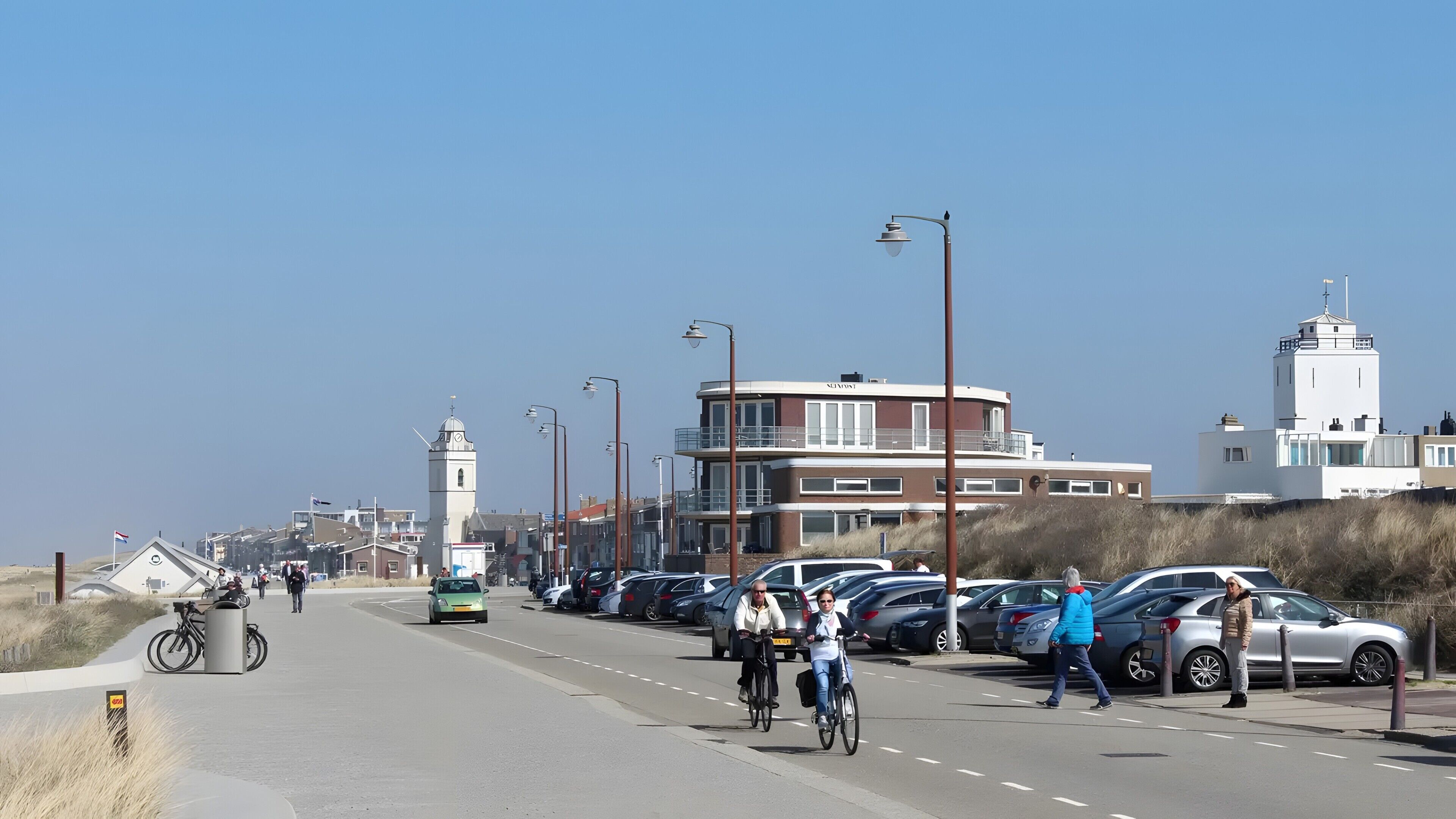 Eine Küstenstraße mit geparkten Autos, Fahrrädern und Fußgängern.