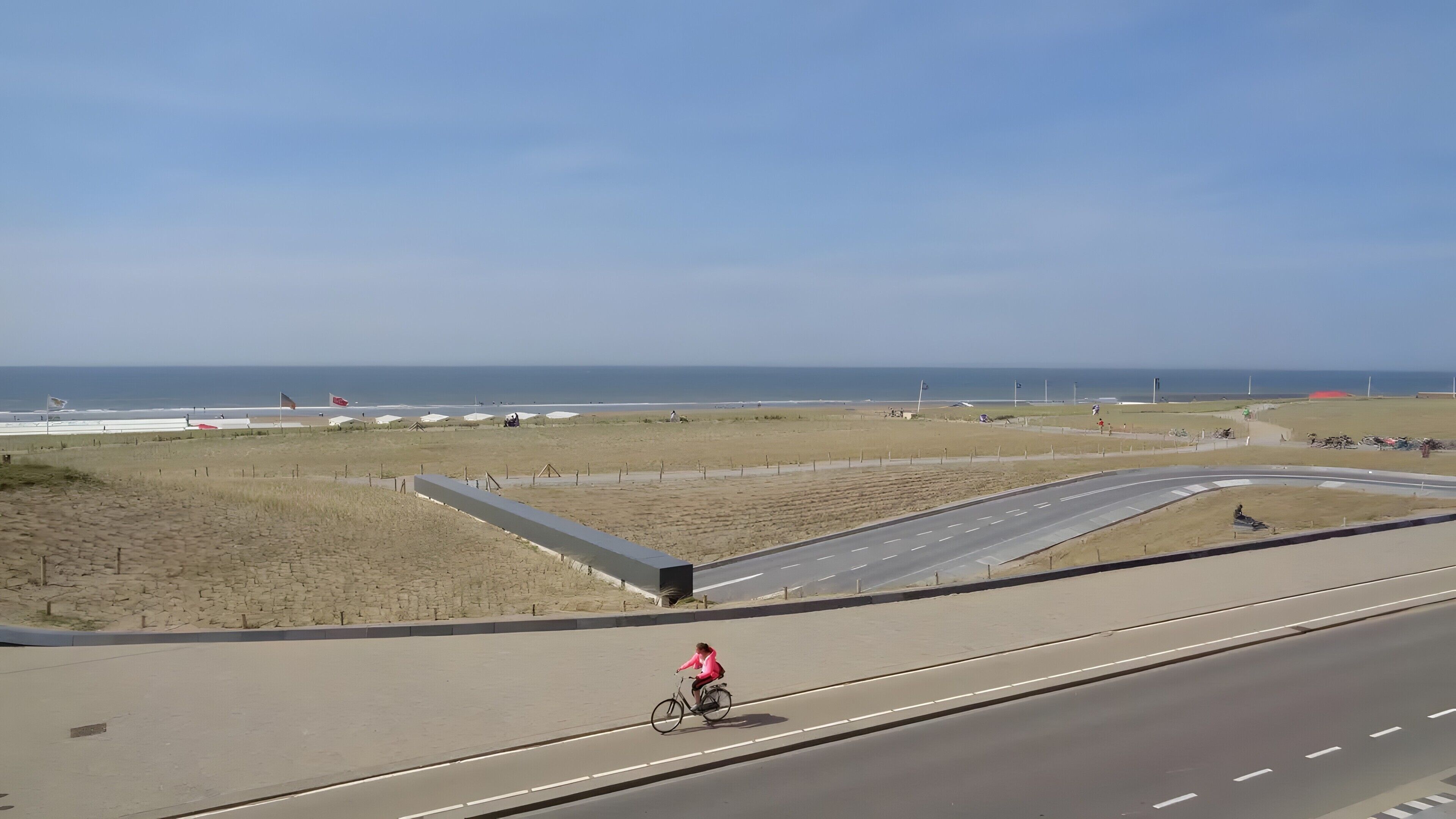 Eine Küstenstraße mit einem Radfahrer, Strand im Hintergrund und wolkenlosem Himmel.