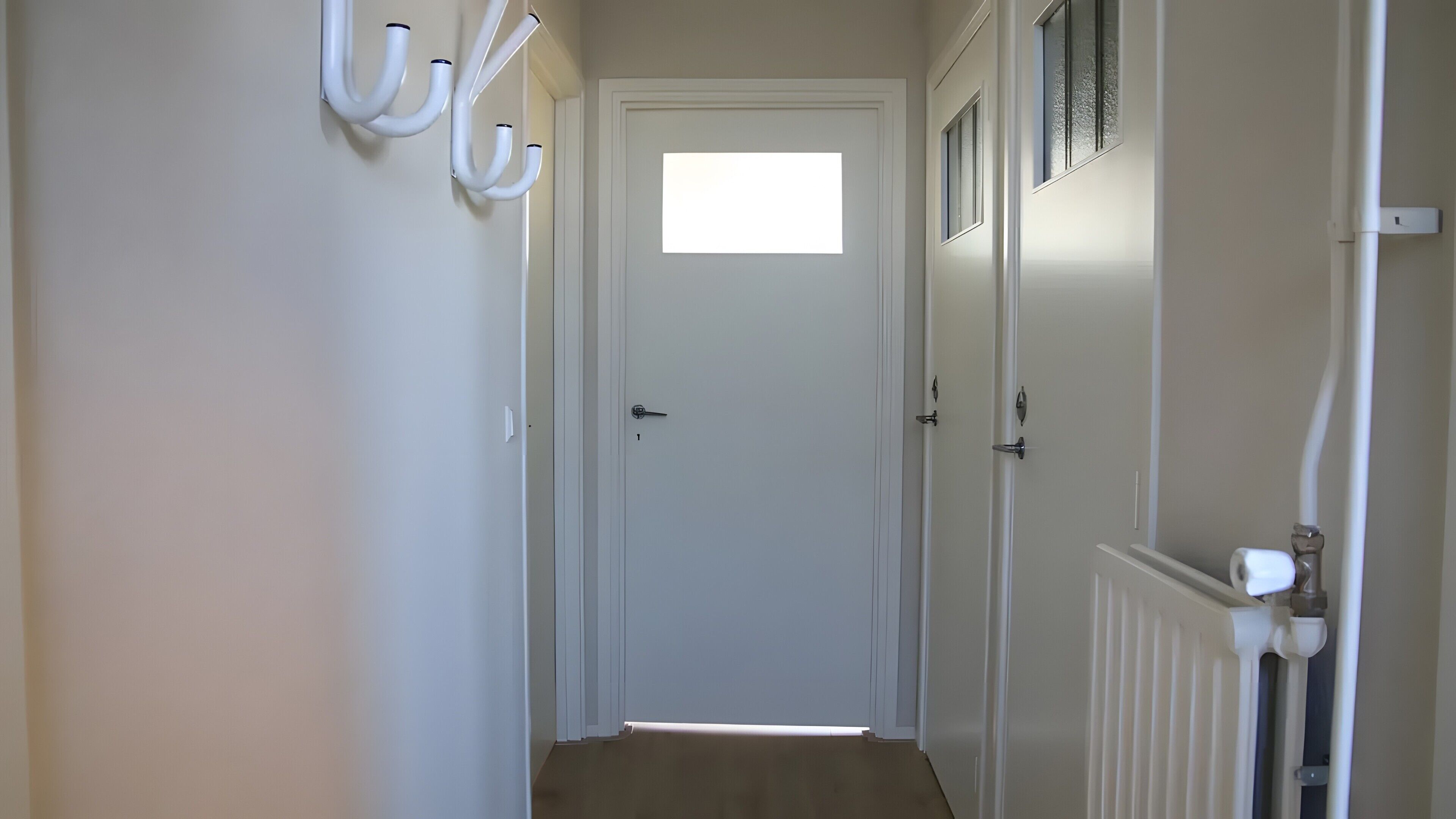 Un pasillo con una puerta blanca, un radiador y una ventana.