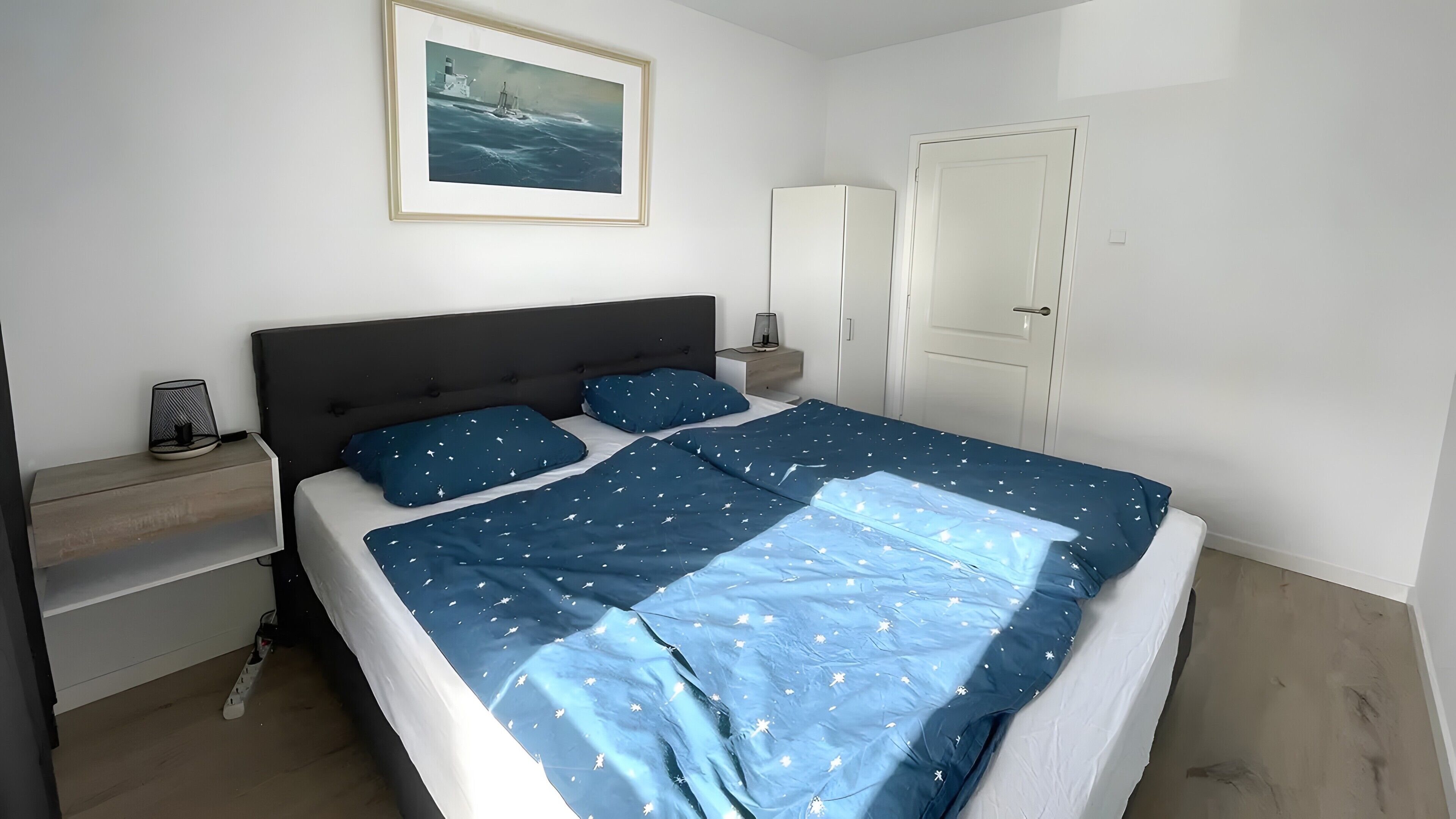 Ein Schlafzimmer mit einem Bett, einem Nachttisch, einer Lampe, einem gerahmten Bild und einer Tür.