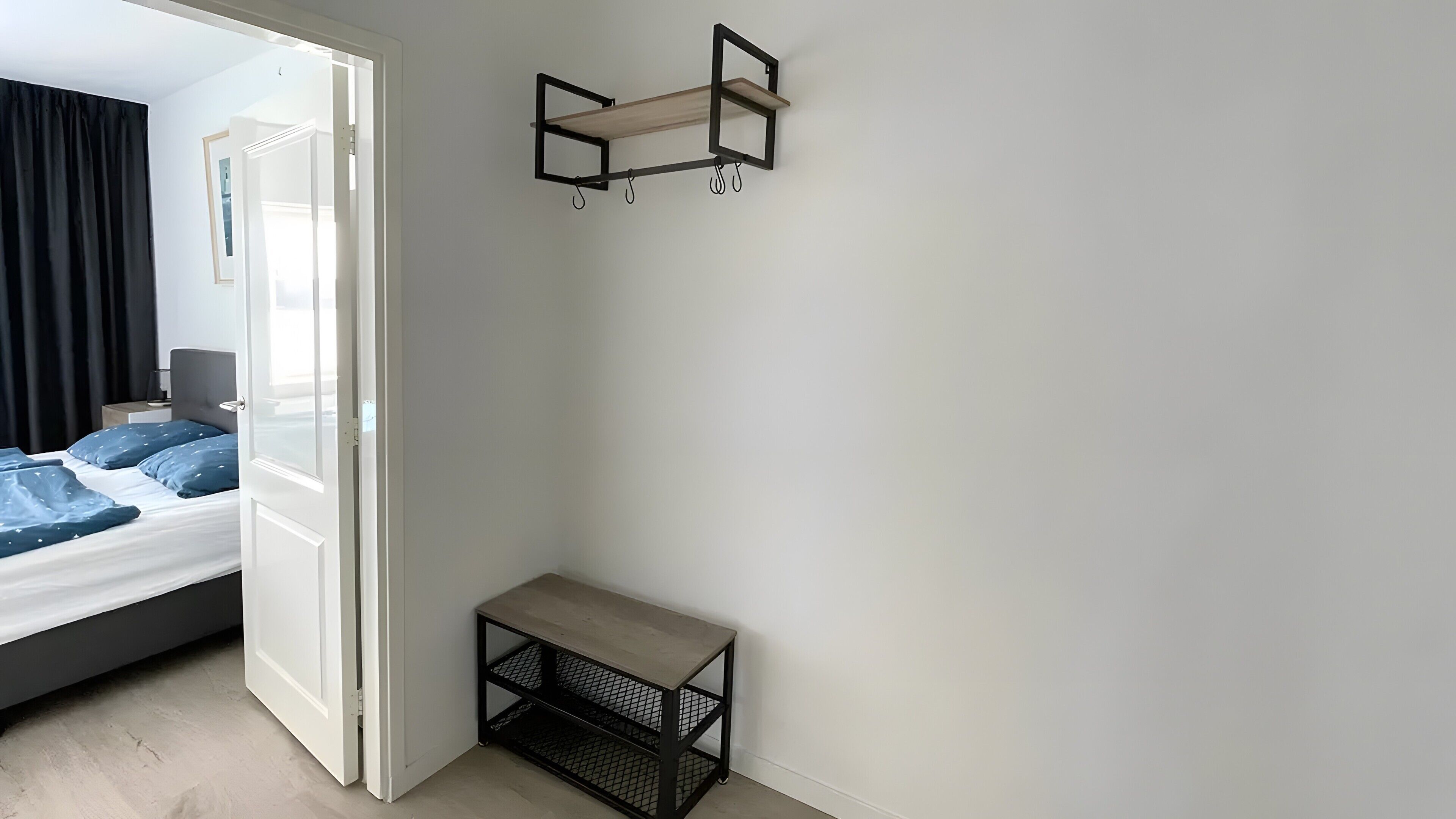 A room with a bed, a small table, a wall-mounted shelf, and a door leading to another room.