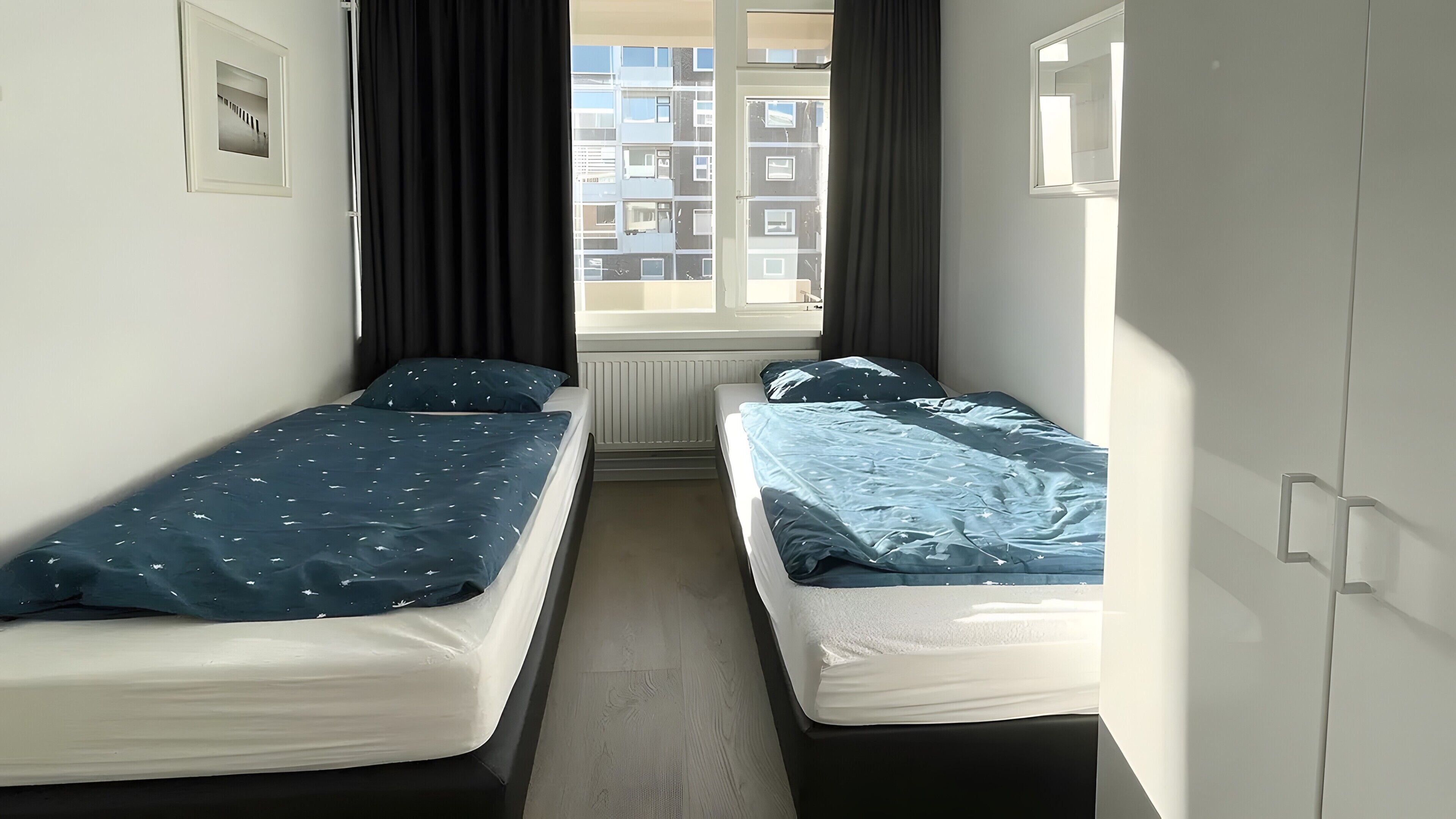 Ein Zimmer mit zwei Einzelbetten, einem fensterbelaubten Fenster, einem Heizkörper und einem Bild an der Wand.