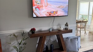 A modern living room with a flat-screen TV mounted on the wall, a wooden coffee table, a potted plant, and a view of the beach through large windows. - Timber Time (Havelock)
