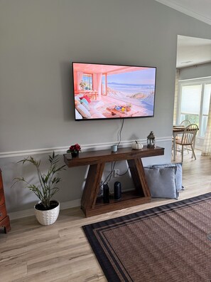 Ein modernes Wohnzimmer mit einem an der Wand befestigten Flachbildfernseher, einem Holztisch, einer Topfpflanze und Blick auf den Strand durch große Fenster.