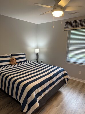 A bedroom with a large bed, a ceiling fan, a bedside table with a lamp, and a window with blinds. - Timber Time (Havelock)