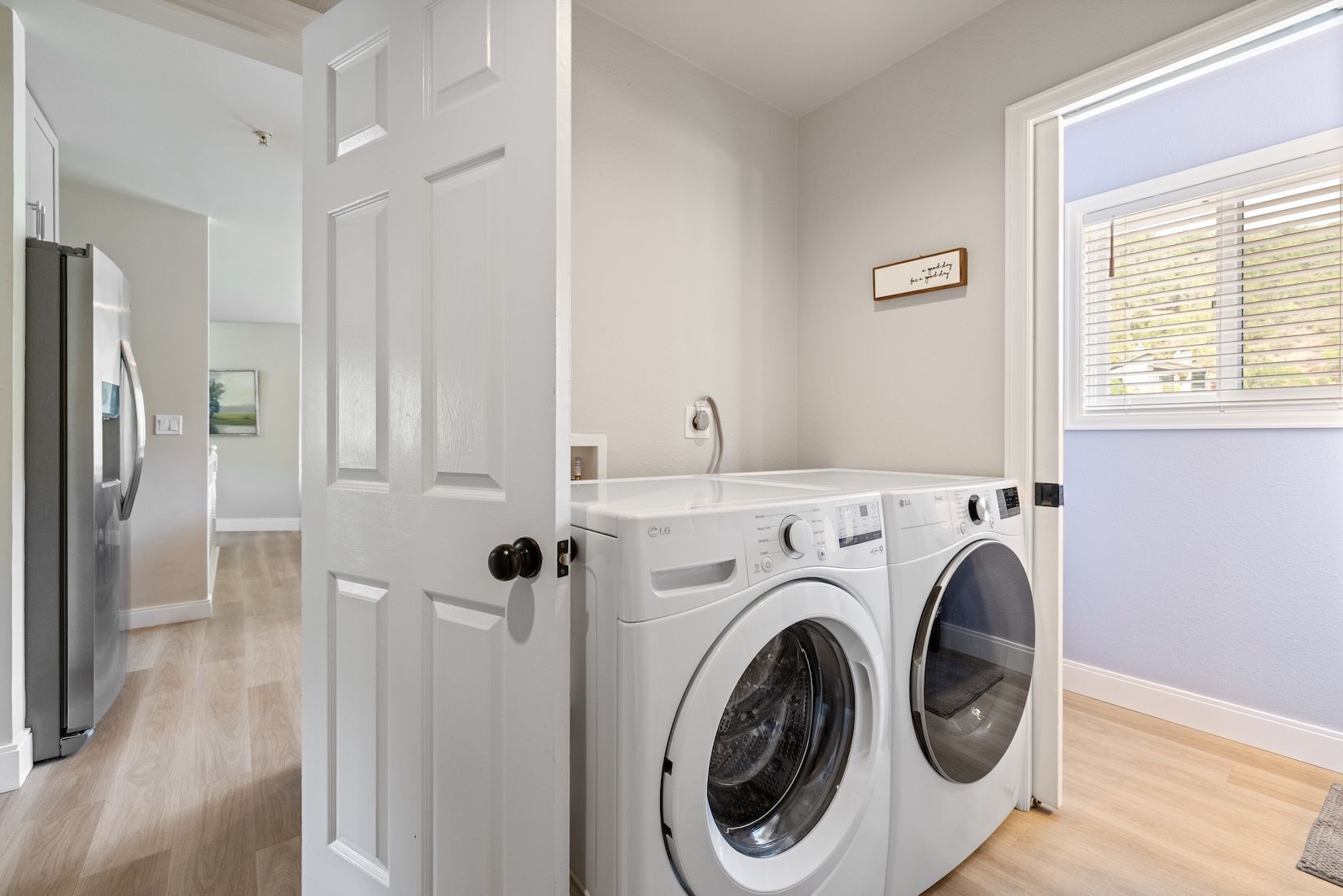 Une buanderie équipée d’un lave-linge et d’un sèche-linge, une porte donnant sur une autre pièce et une fenêtre avec des stores.
