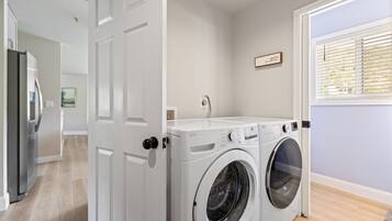 Une buanderie équipée d’un lave-linge et d’un sèche-linge, une porte donnant sur une autre pièce et une fenêtre avec des stores.