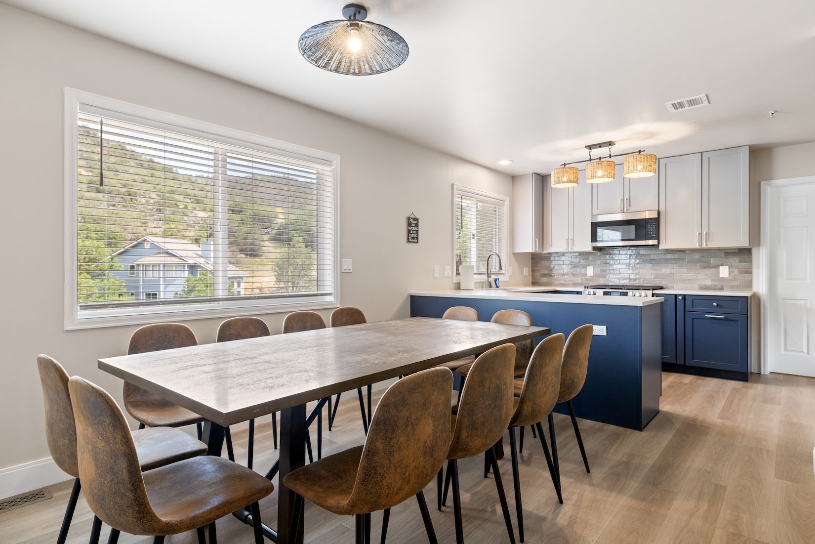 Une cuisine moderne dotée d’un îlot central, d’un coin repas avec une table et des chaises en bois, et de grandes fenêtres équipées de stores.